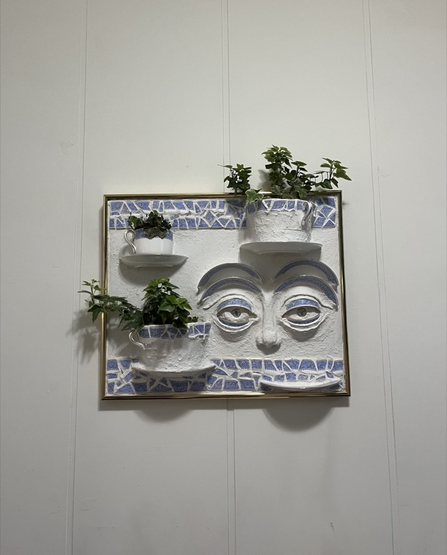 Steeped — a face in white relief with blue mosaic, teacup planters, and trailing ivy